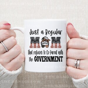 K&ouml;nnte beinhalten: Eine wei&szlig;e Keramiktasse mit einem rot-wei&szlig;-blauen amerikanischen Flaggenmuster. Die Tasse tr&auml;gt den Text "Just a Regular MOM", wobei das "M" durch eine amerikanische Flagge ersetzt wurde. Der Text "That refuses to Co-Parent with The GOVERNMENT" ist unter dem Flaggenmuster aufgedruckt.