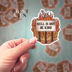 May include: A sticker featuring a sign that reads "HELL IS HOT BE KIND" surrounded by flames. A cartoon devil with a pitchfork sits atop the sign. The sticker has a white border and the text "the BRENT & JEN show" is visible.