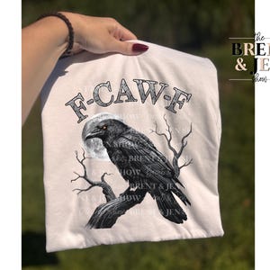 May include: A cream-colored t-shirt featuring a black raven perched on a tree branch in front of a full moon. The text "F-CAW-F" is arched above the raven. The Brent & Jen Show logo is on the right side.