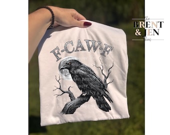 May include: A cream-colored t-shirt featuring a black raven perched on a tree branch in front of a full moon. The text "F-CAW-F" is arched above the raven. The Brent & Jen Show logo is on the right side.