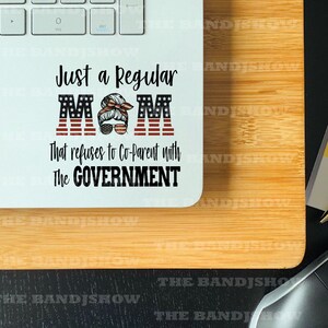 K&ouml;nnte beinhalten: Ein wei&szlig;er Laptop mit einem Aufkleber, auf dem steht "Just a Regular MOM that refuses to Co-Parent with The GOVERNMENT". Der Aufkleber zeigt eine Frau mit einem rot-wei&szlig;-blauen Bandana und Sonnenbrille. Die Buchstaben des Wortes "MOM" sind mit der amerikanischen Flagge verziert.