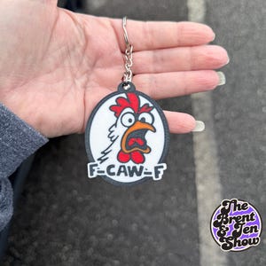 May include: A black and white keychain with a cartoon chicken design. The chicken has a red comb and wattle, an orange beak, and the text "F-CAW-F" below. The keychain is attached to a silver chain.