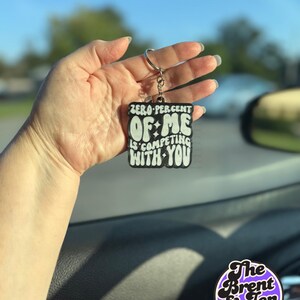 May include: A black and white keychain with the text "ZERO PERCENT OF ME IS COMPETING WITH YOU". The keychain is held up in front of a blurred background. The keychain has a silver key ring.