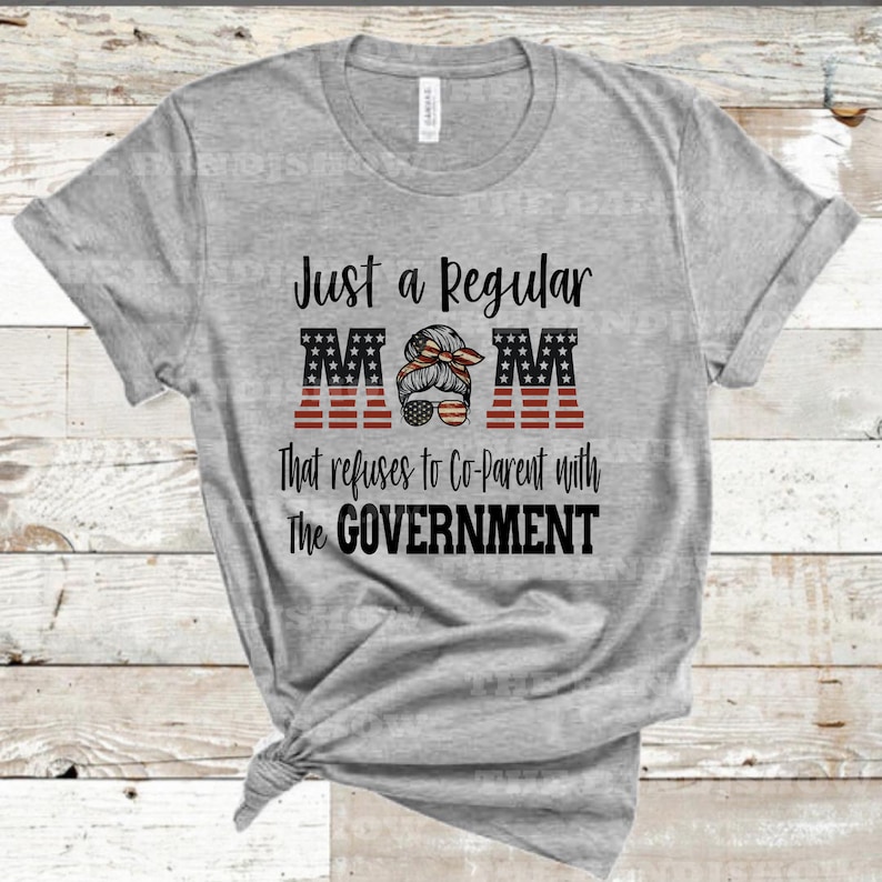 K&ouml;nnte beinhalten: Graues T-Shirt mit dem Text "Just a Regular MOM that refuses to Co-Parent with the GOVERNMENT". Das Wort "MOM" ist in gro&szlig;en Buchstaben mit einem amerikanischen Flaggen-Design. Ein Frauen Gesicht mit einem unordentlichen Dutt und Sonnenbrille befindet sich in der Mitte des Wortes "MOM".