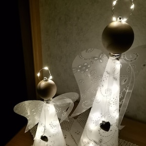 Könnte beinhalten: Zwei weiße, kegelförmige, dekorative Engel mit weißen Flügeln und einer Lichterkette im Inneren. Die Engel stehen auf einer weißen Häkeldeckchen.