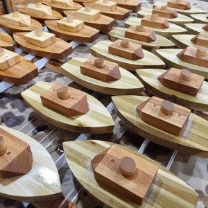 May include: A display of wooden toy boats, each with a light-coloured hull and darker wood cabin and chimney. The boats are arranged on a light-coloured surface. The boats are made of wood.