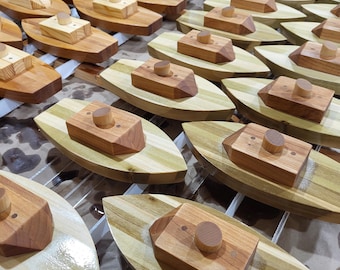May include: A collection of handcrafted wooden toy boats. Each boat features a light-colored hull with darker wood accents and a rectangular cabin topped with a circular chimney. The boats are arranged on a light-colored surface.