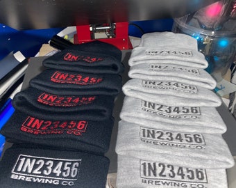 Può includere: Un mucchio di berretti di lana neri e grigi con il logo "IN23456 BREWING CO." ricamato su ciascuno.