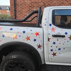 Puede incluir: Una camioneta blanca con una barra antivuelco negra y una caja llena de pegatinas de estrellas de colores. Las estrellas son de varios tonos de azul, rosa y naranja.