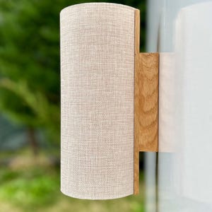 Puede incluir: Aplique de pared cil&iacute;ndrico con pantalla de tela beige y base de madera natural. El aplique est&aacute; montado sobre una superficie blanca, con un fondo verde borroso. Luminaria para iluminaci&oacute;n interior.