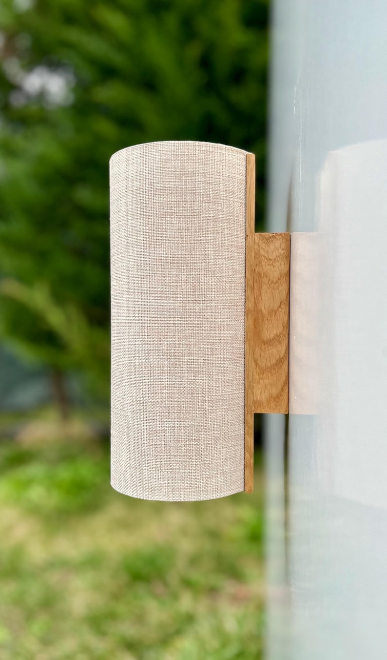 Puede incluir: Aplique de pared cil&iacute;ndrico con pantalla de tela beige y base de madera natural. El aplique est&aacute; montado sobre una superficie blanca, con un fondo verde borroso. Luminaria para iluminaci&oacute;n interior.
