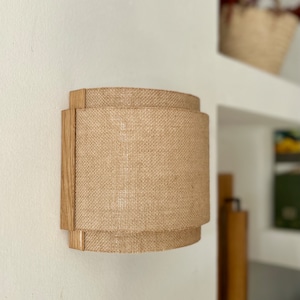 May include: A wall-mounted light fixture with a natural wood frame and a beige burlap shade. The shade is cylindrical and has a slightly curved shape.