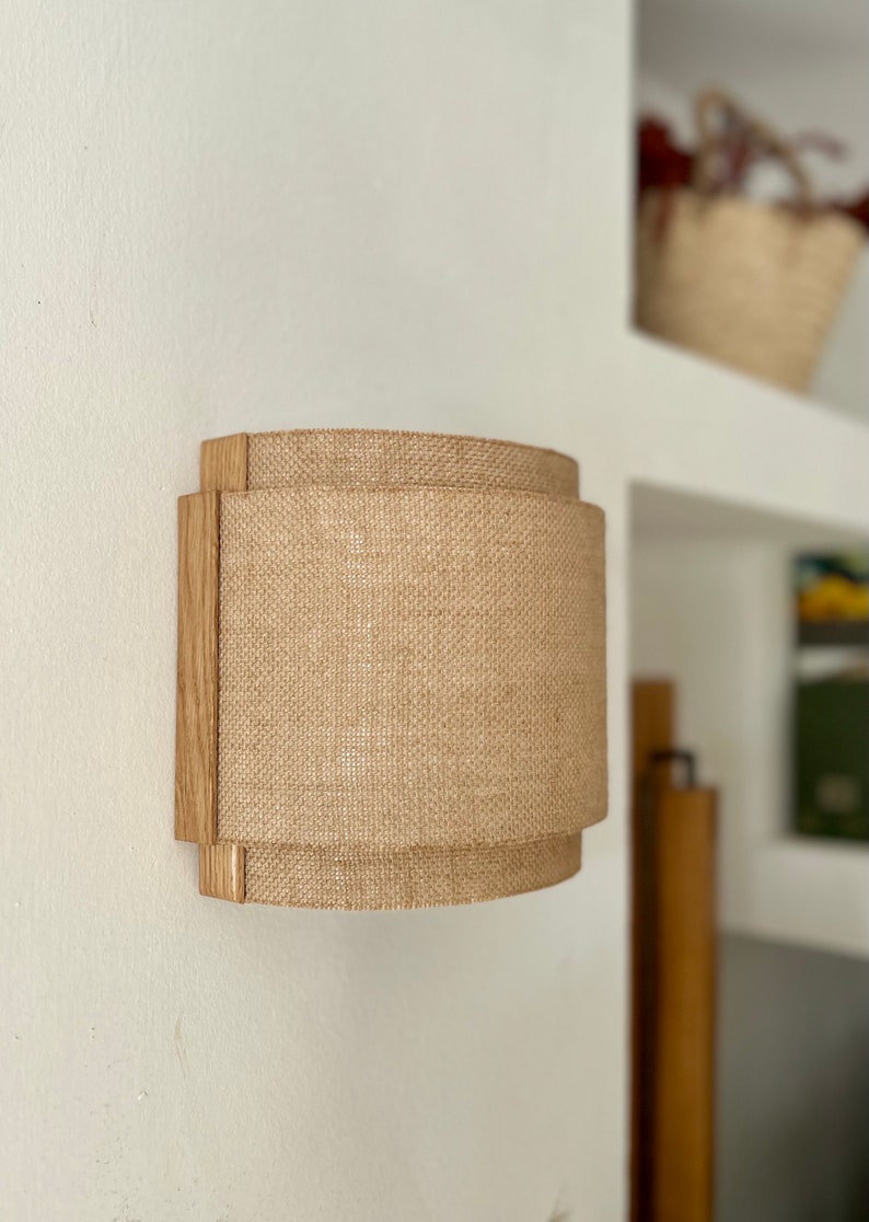 May include: A wall-mounted light fixture with a natural wood frame and a beige burlap shade. The shade is cylindrical and has a slightly curved shape.