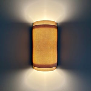 Op de afbeelding: Een cilindrische wandlamp met een getextureerde, lichtbruine kap, omlijst door een donkerbruine rand. Het licht straalt een warme gloed uit, met licht dat naar boven en beneden schijnt. De armatuur is op een muur gemonteerd.