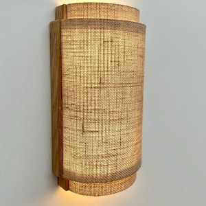 May include: A wall-mounted light fixture with a natural wood frame and a woven fabric shade. The shade is a light beige color and has a slightly curved shape.
