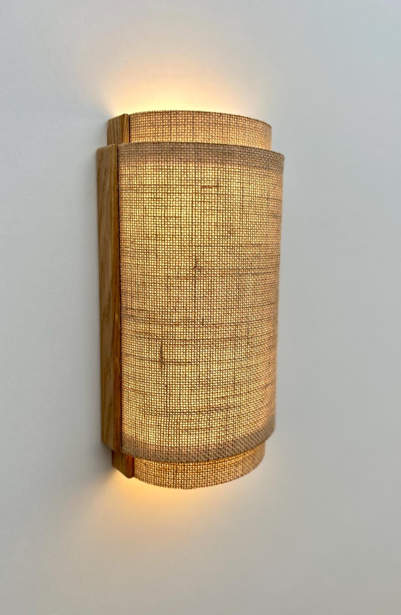 May include: A wall-mounted light fixture with a natural wood frame and a woven fabric shade. The shade is a light beige color and has a slightly curved shape.