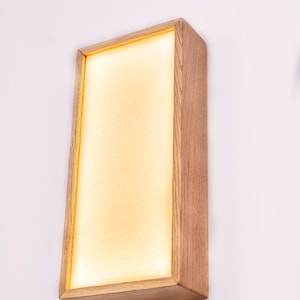 Pu&ograve; includere: Un'applique da parete rettangolare in legno con una luce bianca calda che emana dal centro. L'applique ha una finitura in legno naturale e un design semplice e moderno.