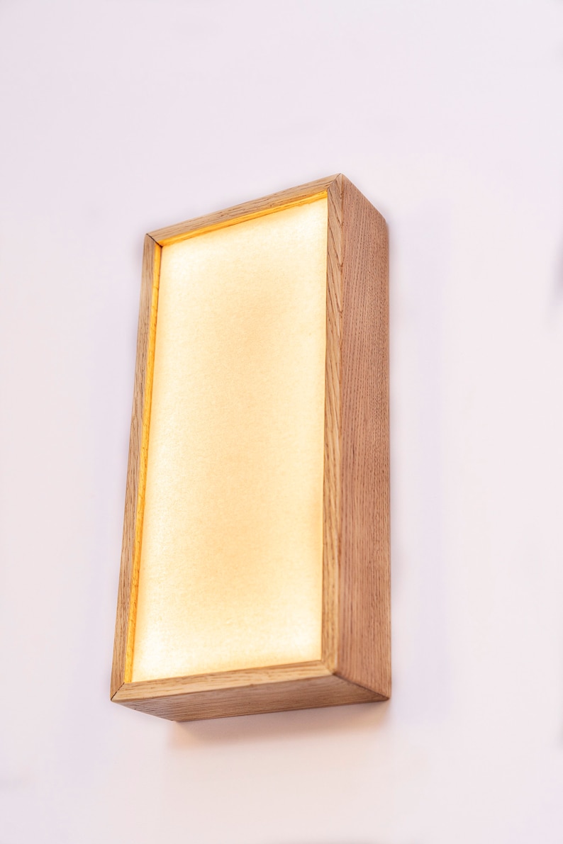 Pu&ograve; includere: Un'applique da parete rettangolare in legno con una luce bianca calda che emana dal centro. L'applique ha una finitura in legno naturale e un design semplice e moderno.