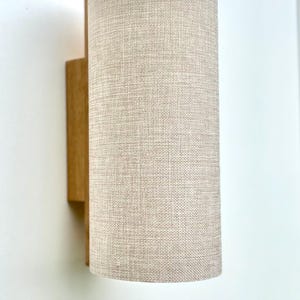 Puede incluir: Aplique de pared cil&iacute;ndrico con pantalla de tela beige y base de madera. La pantalla tiene un aspecto texturizado y la base de madera es rectangular. El aplique est&aacute; montado en una pared blanca.