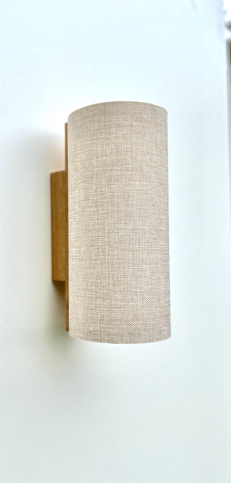 Puede incluir: Aplique de pared cil&iacute;ndrico con pantalla de tela beige y base de madera. La pantalla tiene un aspecto texturizado y la base de madera es rectangular. El aplique est&aacute; montado en una pared blanca.