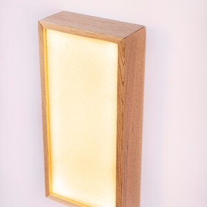 Pu&ograve; includere: Un'applique da parete rettangolare con una cornice in legno chiaro e un pannello bianco illuminato. L'applique &egrave; montata su una parete bianca.