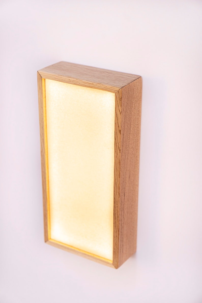 Pu&ograve; includere: Un'applique da parete rettangolare con una cornice in legno chiaro e un pannello bianco illuminato. L'applique &egrave; montata su una parete bianca.