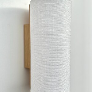 May include: A white fabric lampshade with a wood wall mount. The lampshade has a textured woven pattern.