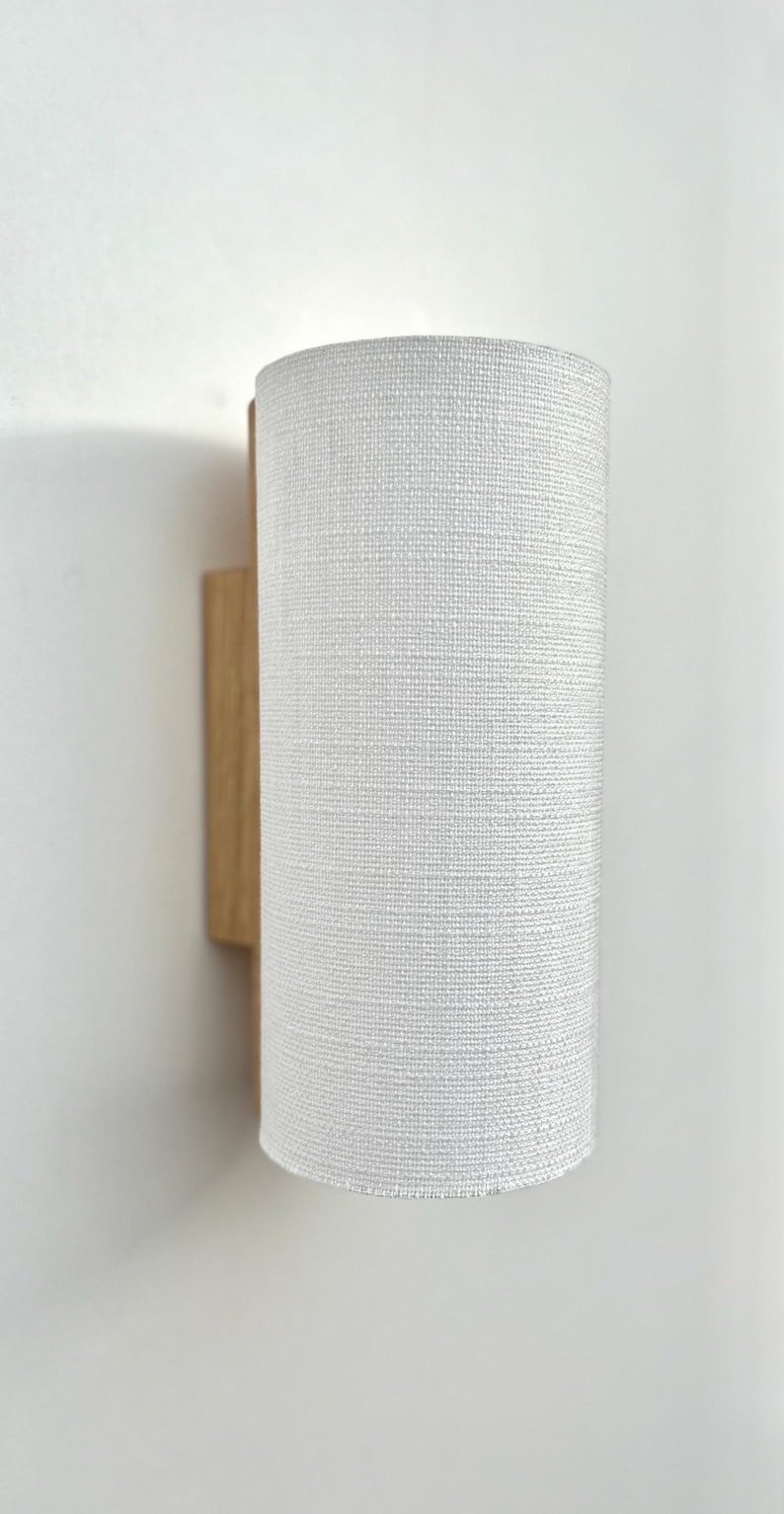 May include: A white fabric lampshade with a wood wall mount. The lampshade has a textured woven pattern.