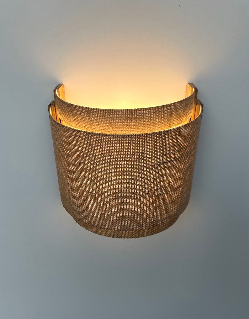 May include: A wall-mounted light fixture with a woven fabric shade. The shade is a light brown color and has a slightly curved shape. The light is turned on and illuminating the fabric.