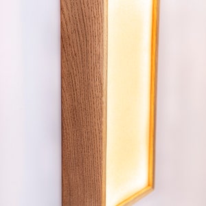 Pu&ograve; includere: Un'applique da parete rettangolare con una cornice in legno chiaro e un pannello bianco illuminato. L'applique &egrave; montata su una parete bianca.