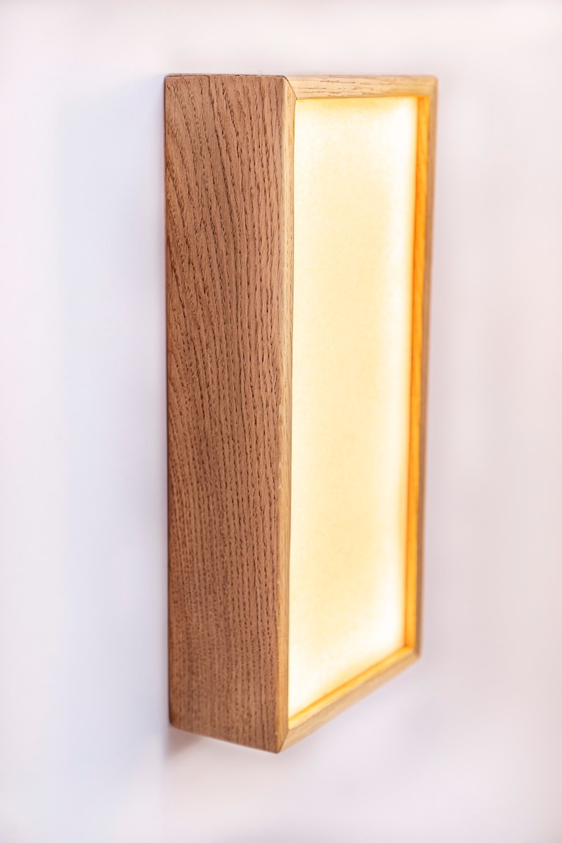 Pu&ograve; includere: Un'applique da parete rettangolare con una cornice in legno chiaro e un pannello bianco illuminato. L'applique &egrave; montata su una parete bianca.