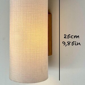 May include: A white fabric cylindrical wall lamp with a wooden base. The lamp is 9.8 inches tall and 4.7 inches wide.