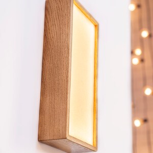 Pu&ograve; includere: Un'applique da parete rettangolare in legno con una luce all'interno. L'applique &egrave; montata su una parete bianca e ha un bagliore caldo e invitante.