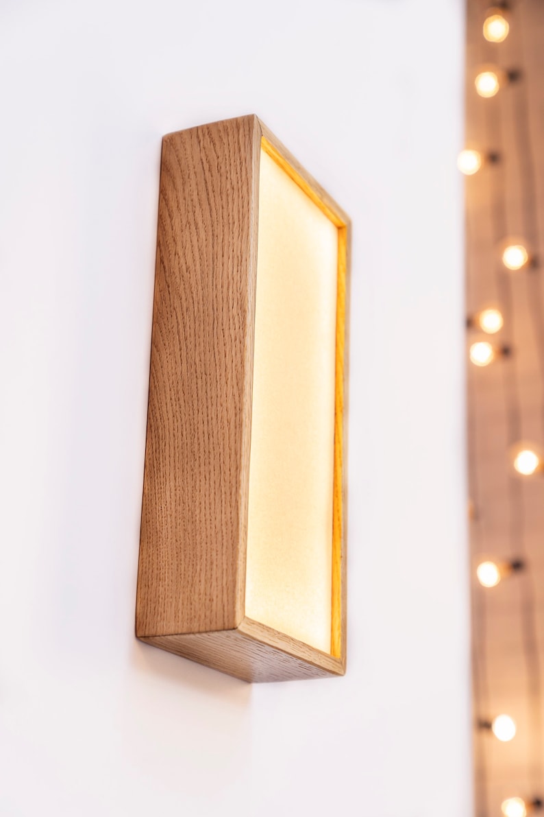 Pu&ograve; includere: Un'applique da parete rettangolare in legno con una luce all'interno. L'applique &egrave; montata su una parete bianca e ha un bagliore caldo e invitante.