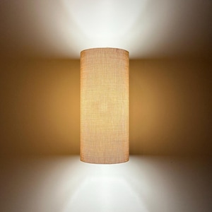 May include: A cylindrical wall sconce with a textured, beige fabric shade. The light fixture is mounted on a neutral-toned wall, casting a warm glow above and below. The design is simple and modern, suitable for various interior styles.