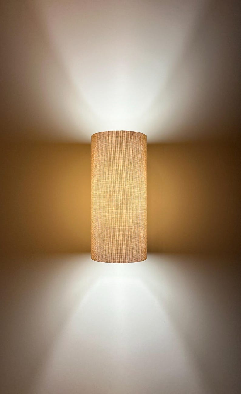 Puede incluir: Aplique de pared cil&iacute;ndrico con pantalla de tela beige texturizada. La l&aacute;mpara est&aacute; montada en una pared de tono neutro, proyectando un brillo c&aacute;lido arriba y abajo. Dise&ntilde;o sencillo y moderno, adecuado para varios estilos de interior.