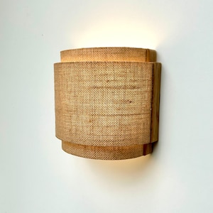 May include: A wall-mounted light fixture with a natural woven fabric shade and a wooden frame. The light is turned on and illuminating the fabric shade.