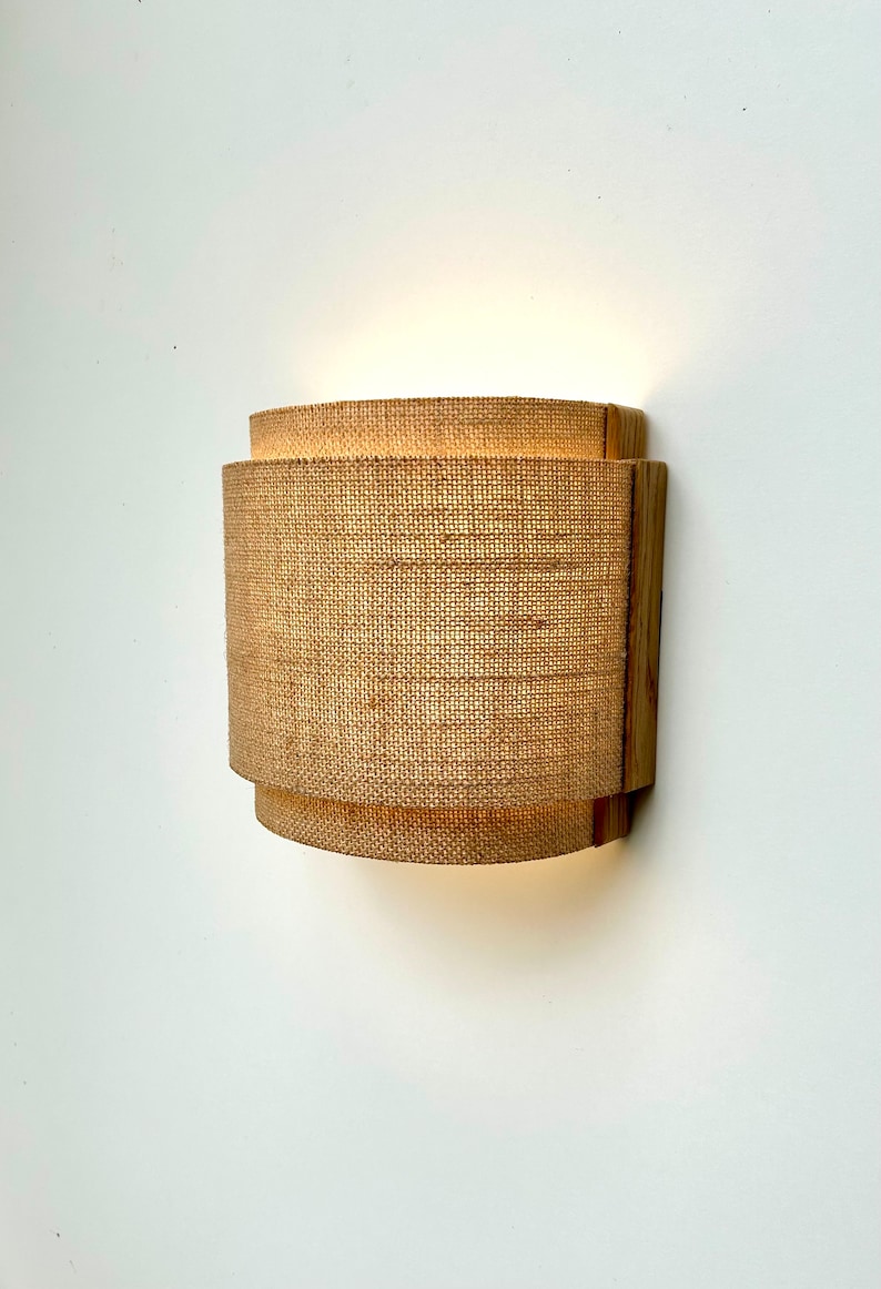May include: A wall-mounted light fixture with a natural woven fabric shade and a wooden frame. The light is turned on and illuminating the fabric shade.