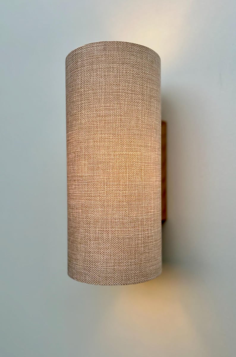 Puede incluir: Aplique de pared cil&iacute;ndrico con pantalla de tela beige. La pantalla tiene un aspecto texturizado y est&aacute; montada sobre una base de madera. El aplique est&aacute; dise&ntilde;ado para iluminaci&oacute;n interior.