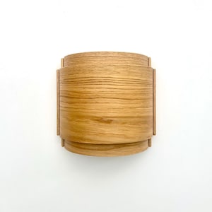 May include: A light brown, wooden wall sconce with a curved, layered design. The sconce has a rectangular shape with rounded edges and a natural wood grain finish. The design is modern and minimalist.