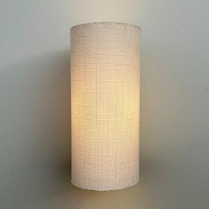 May include: A white cylindrical lampshade with a woven texture. The lampshade is lit and casts a warm glow.