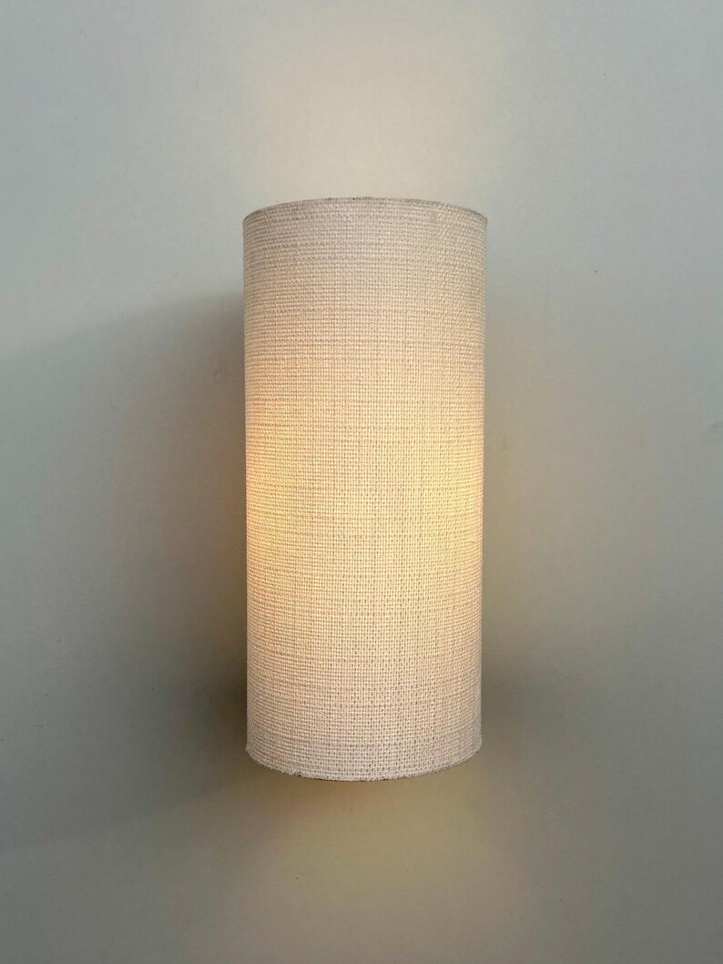 May include: A white cylindrical lampshade with a woven texture. The lampshade is lit and casts a warm glow.