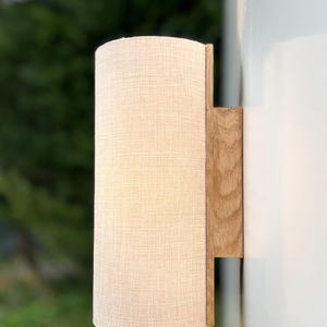 Puede incluir: Una l&aacute;mpara de pared con una pantalla cil&iacute;ndrica de tela blanquecina. La pantalla est&aacute; unida a una base rectangular de madera clara. La l&aacute;mpara est&aacute; montada en una pared blanca, con un fondo verde borroso.