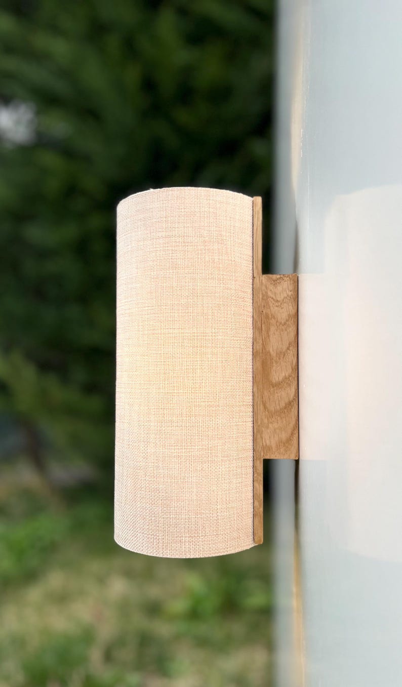 Puede incluir: Una l&aacute;mpara de pared con una pantalla cil&iacute;ndrica de tela blanquecina. La pantalla est&aacute; unida a una base rectangular de madera clara. La l&aacute;mpara est&aacute; montada en una pared blanca, con un fondo verde borroso.