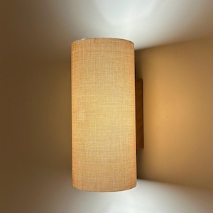 Puede incluir: Aplique de pared cil&iacute;ndrico con pantalla de tela beige texturizada. El aplique est&aacute; montado sobre un respaldo de madera y emite una luz c&aacute;lida, creando un ambiente acogedor. Dise&ntilde;o sencillo y moderno.