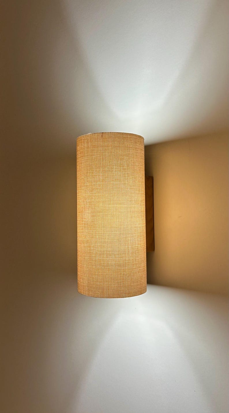 Puede incluir: Aplique de pared cil&iacute;ndrico con pantalla de tela beige texturizada. El aplique est&aacute; montado sobre un respaldo de madera y emite una luz c&aacute;lida, creando un ambiente acogedor. Dise&ntilde;o sencillo y moderno.