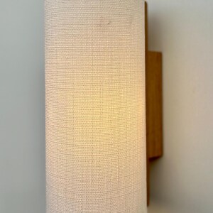 May include: A white fabric lampshade with a wooden wall mount. The lampshade is cylindrical and has a textured surface.