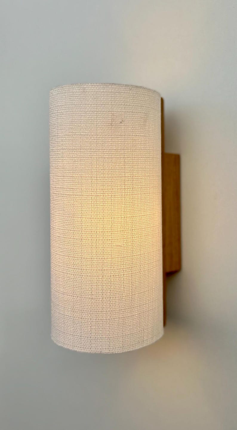 May include: A white fabric lampshade with a wooden wall mount. The lampshade is cylindrical and has a textured surface.