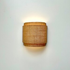 May include: A wall-mounted light fixture with a woven, cylindrical shade. The shade is made of a natural material, like jute or hemp, and has a warm, inviting glow.