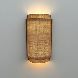 May include: A wall-mounted light fixture with a woven fabric shade. The shade is a light brown color and has a rectangular shape. The light is turned on and illuminating the room.